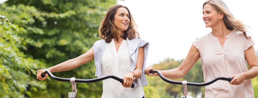 Two smiling women walking outside with bicycles, happy that they decided to get varicose vein treatment